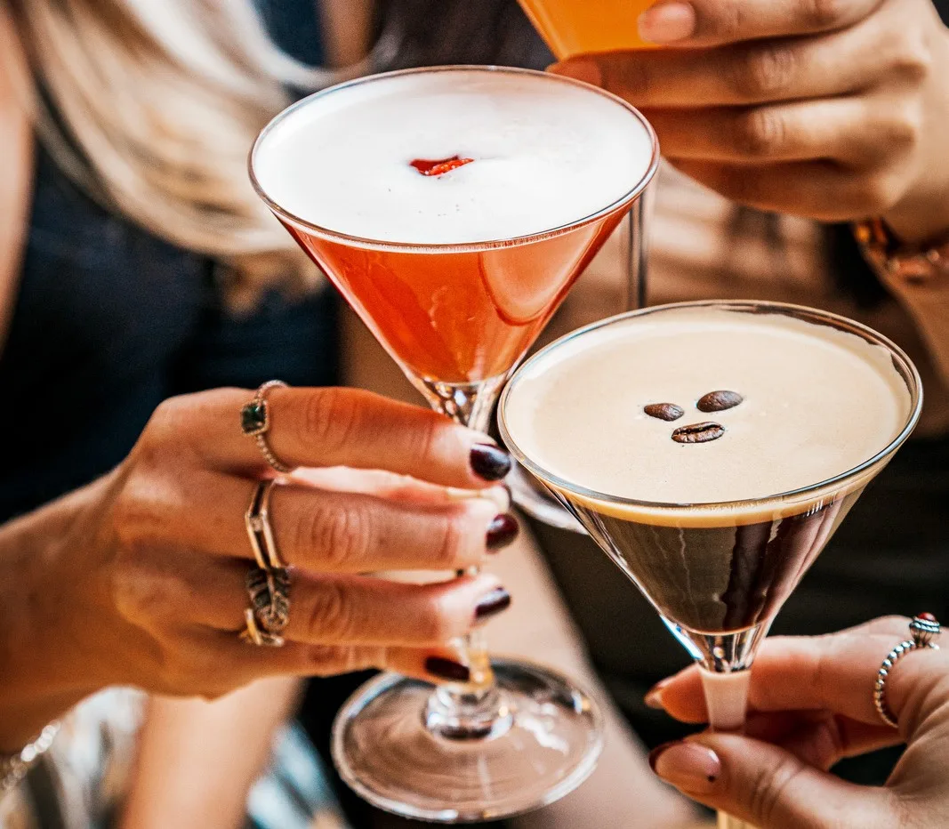 Two women clinking cocktail glasses. One cocktail look fruity whilst the other is coffee flavoured.