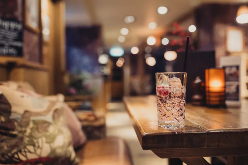 A blurred picture of a pub. In focus there is a drink on the table with a raspberry in it and a straw coming out of it