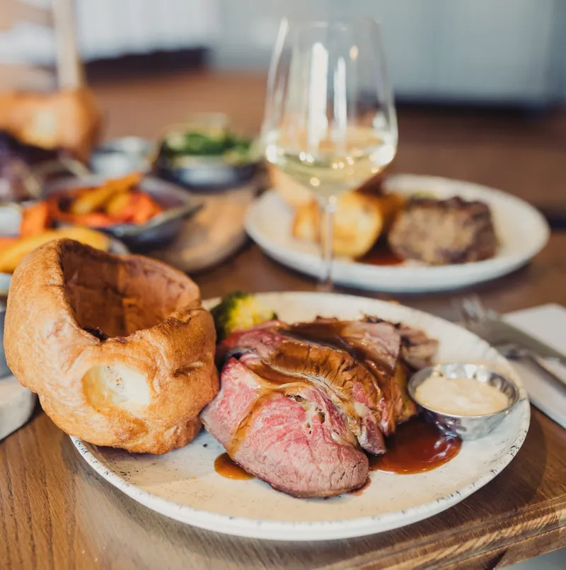 A plate with yorskshire pudding and beef. Behind the plate is a wine in a glass.