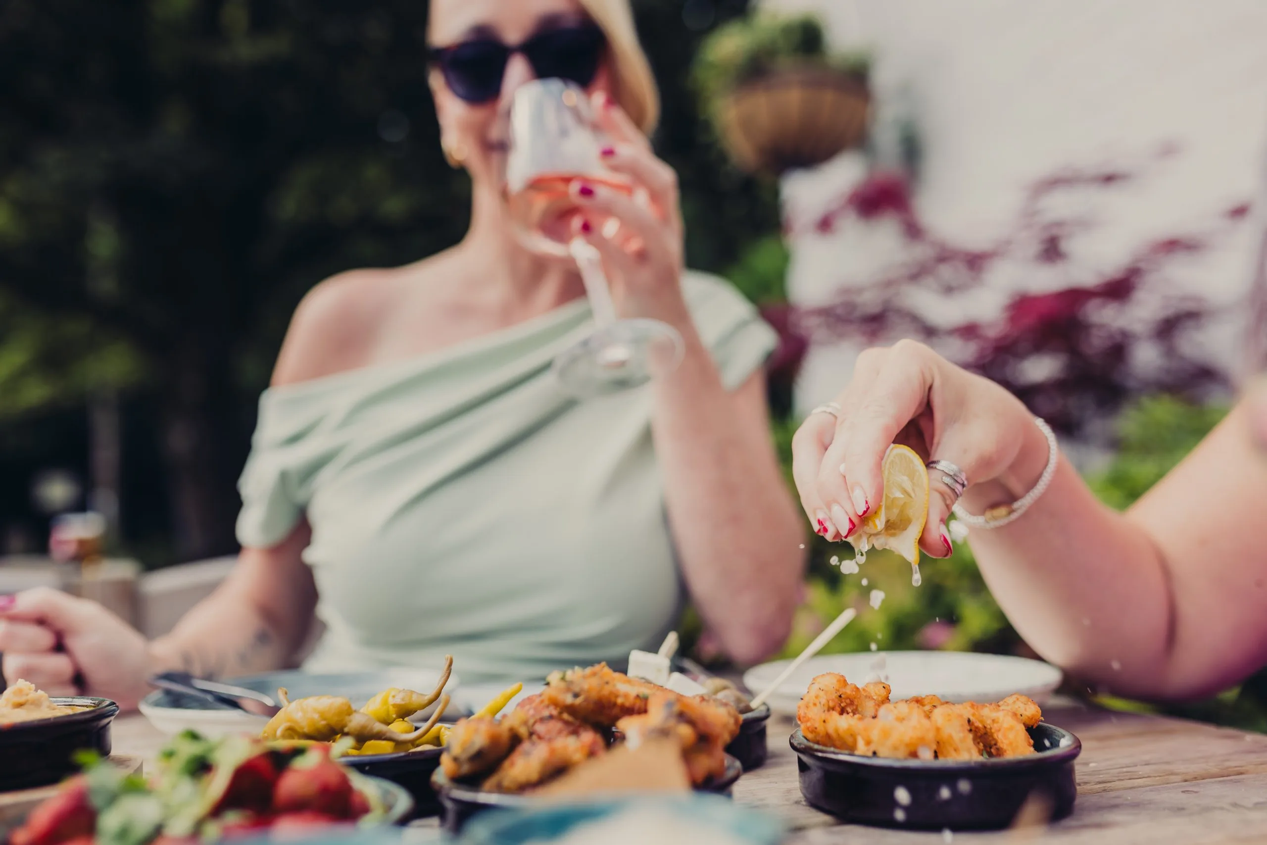 Several dishes on a table. Someone is squeezing a lemon juice onto one of the dishes whilst a lady in the background is drinking